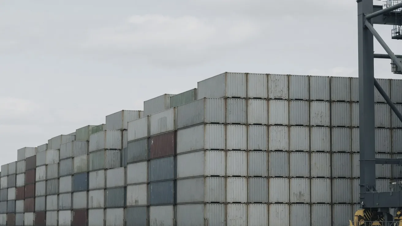 Contenedores apilados en una terminal maritima bajo un cielo nublado, con la pata de una grua de muelle a la derecha.